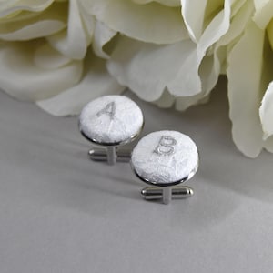 May include: A pair of white lace cufflinks with silver metal clasps. The cufflinks are embroidered with the letters "A" and "B".