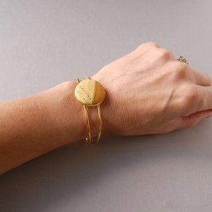 May include: A gold-toned metal cuff bracelet featuring a circular centerpiece. The centerpiece is divided into two sections: one side is a solid mustard yellow fabric, and the other is a lighter yellow fabric with a subtle white dotted pattern.