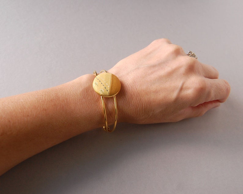 May include: A gold-toned metal cuff bracelet featuring a circular centerpiece. The centerpiece is divided into two sections: one side is a solid mustard yellow fabric, and the other is a lighter yellow fabric with a subtle white dotted pattern.
