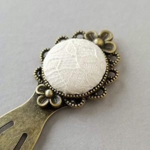 May include: A white lace bookmark with a bronze metal frame and floral accents.