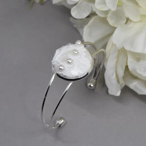 May include: A silver-toned cuff bracelet featuring a circular centerpiece covered in white fabric with a subtle checkered pattern. The center is embellished with delicate white lace trim and three small, round, white faux pearls arranged vertically.