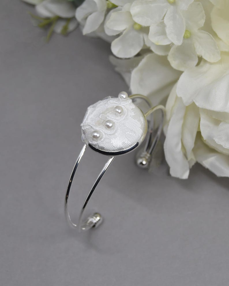 May include: A silver-toned cuff bracelet featuring a circular centerpiece covered in white fabric with a subtle checkered pattern. The center is embellished with delicate white lace trim and three small, round, white faux pearls arranged vertically.