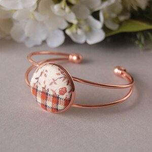 May include: A rose gold-toned metal cuff bracelet featuring a circular centerpiece. The center is inlaid with fabric displaying a split design: the top half has a cream and brown floral pattern, while the bottom half features a brown and cream plaid.