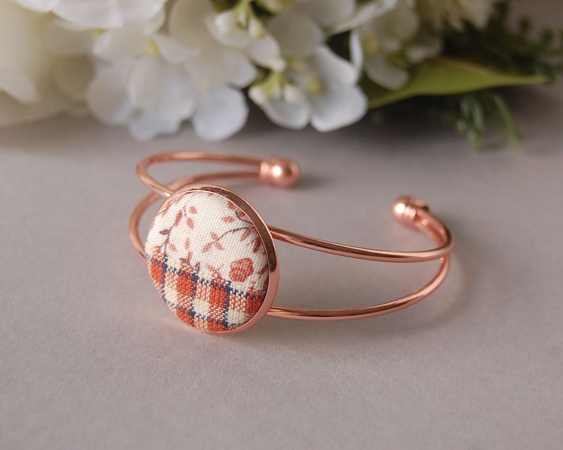 May include: A rose gold-toned metal cuff bracelet featuring a circular centerpiece. The center is inlaid with fabric displaying a split design: the top half has a cream and brown floral pattern, while the bottom half features a brown and cream plaid.