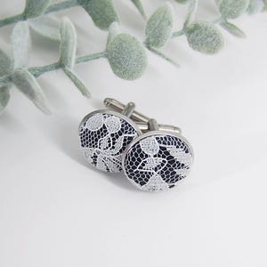 May include: A pair of silver cufflinks with black and white lace designs. The lace is white and the background is black.