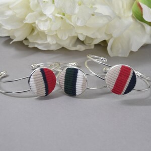 May include: Three silver-toned metal cuff bracelets, each featuring a circular centerpiece covered in ribbed fabric. The fabric discs display vertical stripes in varying color combinations of beige, red, navy blue, white, and dark green.