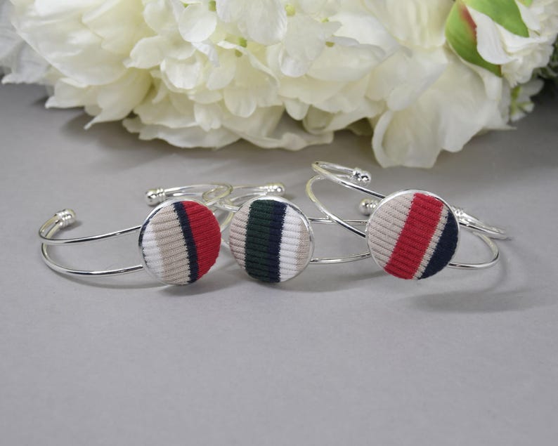May include: Three silver-toned metal cuff bracelets, each featuring a circular centerpiece covered in ribbed fabric. The fabric discs display vertical stripes in varying color combinations of beige, red, navy blue, white, and dark green.