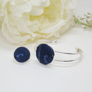May include: A silver-toned cuff bracelet and a matching circular pin, both featuring a dark navy blue fabric inlay with a textured pattern and subtle, shimmering blue sequin accents arranged in a curved line across the surface of each piece.
