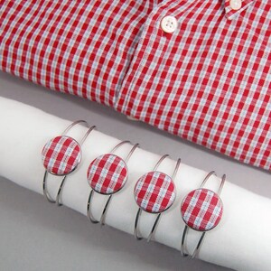 May include: Four silver-toned metal cuff bracelets, each featuring a circular fabric button with a red and white gingham check pattern, displayed on a white cylindrical roll next to a red and white gingham button-down shirt with white buttons.
