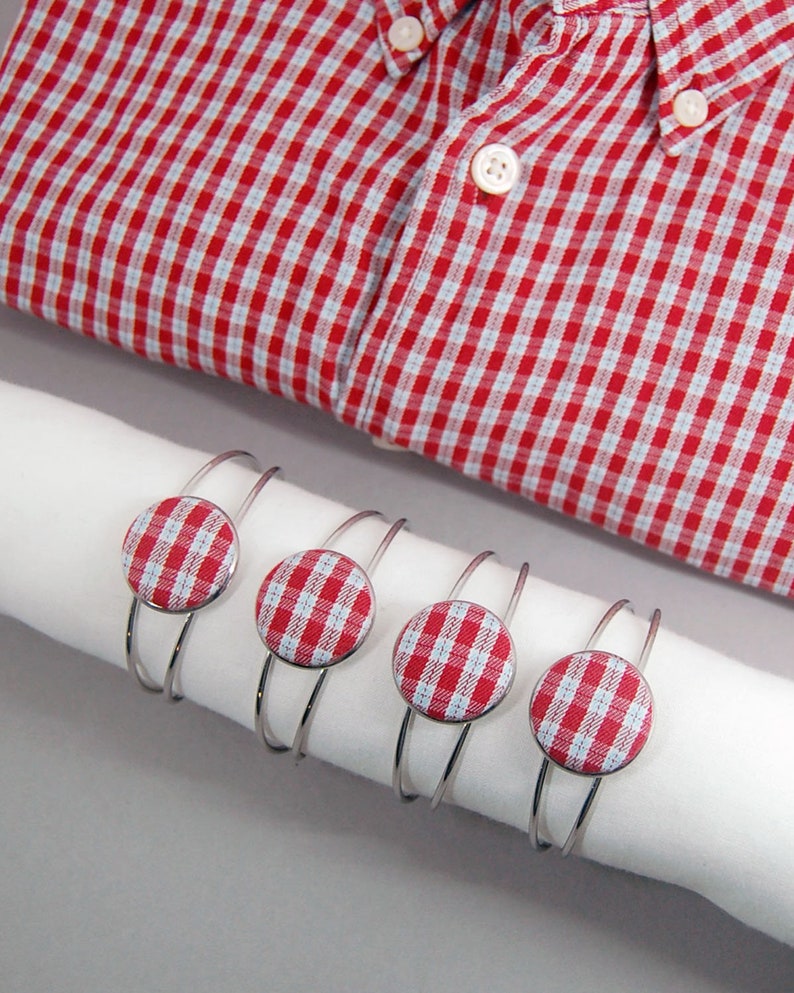 May include: Four silver-toned metal cuff bracelets, each featuring a circular fabric button with a red and white gingham check pattern, displayed on a white cylindrical roll next to a red and white gingham button-down shirt with white buttons.