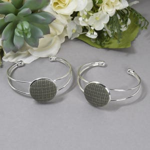 May include: Two silver-toned cuff bracelets featuring a double-band design and a circular centerpiece. Each centerpiece is covered in a textured, olive-green fabric with a subtle, light-colored grid pattern across the surface.