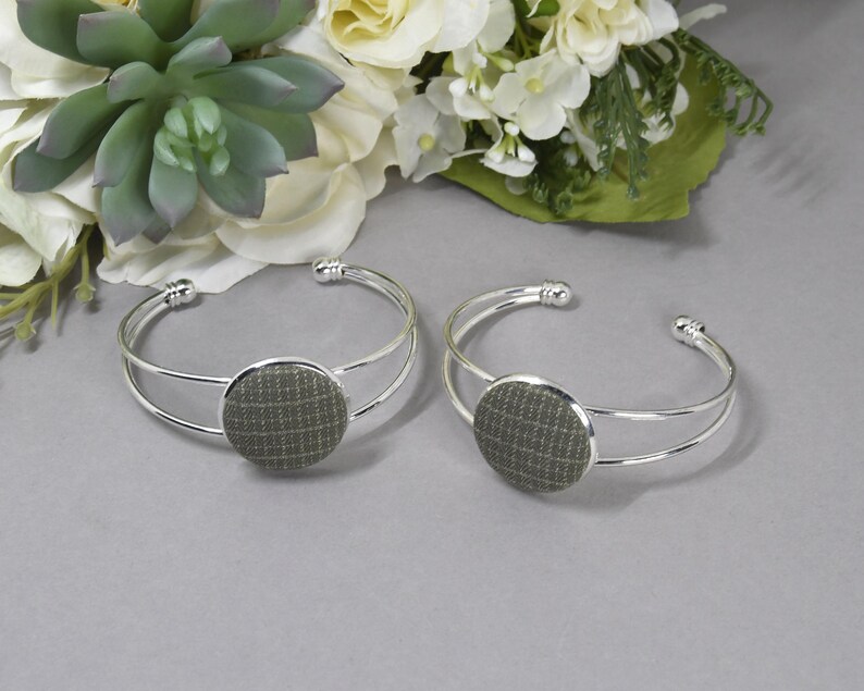 May include: Two silver-toned cuff bracelets featuring a double-band design and a circular centerpiece. Each centerpiece is covered in a textured, olive-green fabric with a subtle, light-colored grid pattern across the surface.