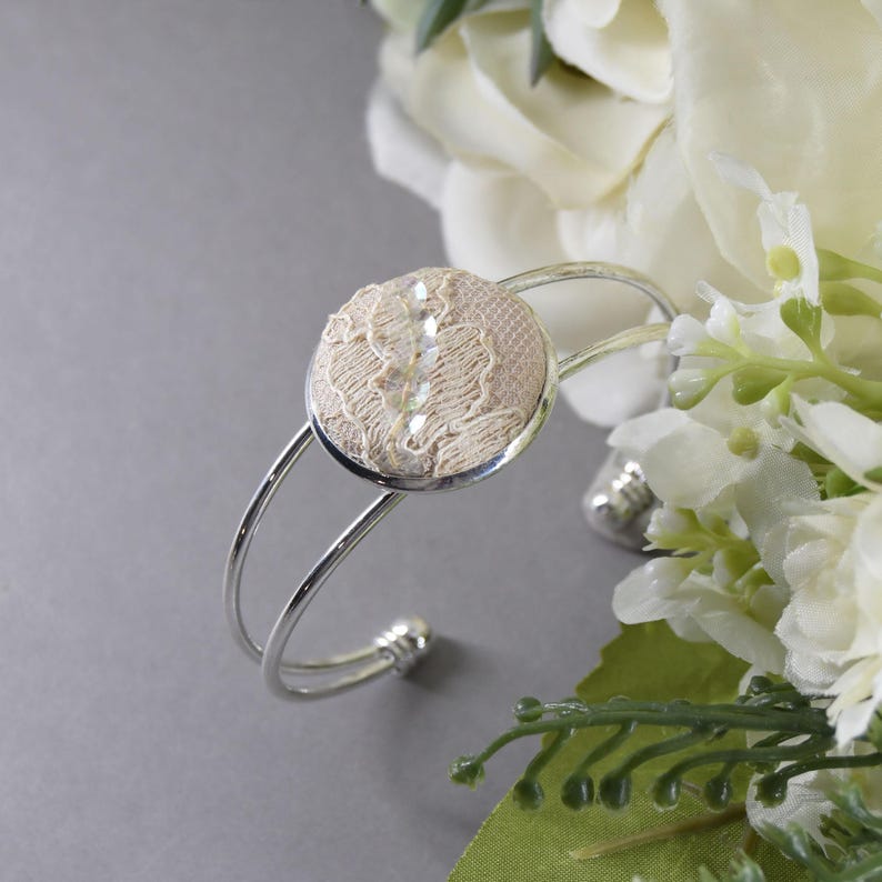 May include: A silver-toned metal cuff bracelet featuring a circular centerpiece. The center is inlaid with beige lace fabric and a vertical row of clear, faceted crystals. The bracelet has a double-band design that remains open at the back.