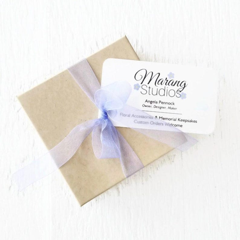 May include: A square brown cardboard gift box tied with a light purple sheer organza ribbon bow. A white rectangular business card is tucked under the ribbon, featuring the text Marang Studios, Angela Pennock, and Floral Accessories & Memorial Keepsakes.