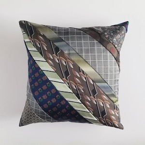 May include: Square decorative pillow made from repurposed neckties. The pillow features a patchwork design with diagonal stripes of various colors and patterns, including navy, green, brown, and gray. The pillow is a unique home decor item.