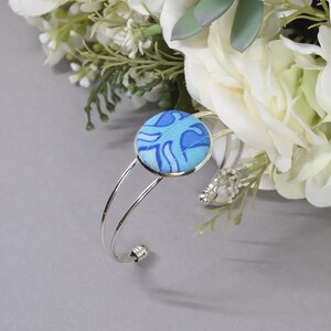 May include: A silver-toned metal cuff bracelet featuring a circular centerpiece covered in light and dark blue fabric with a swirling pattern. The bracelet has a double-wire band design that terminates in small, rounded decorative end caps.
