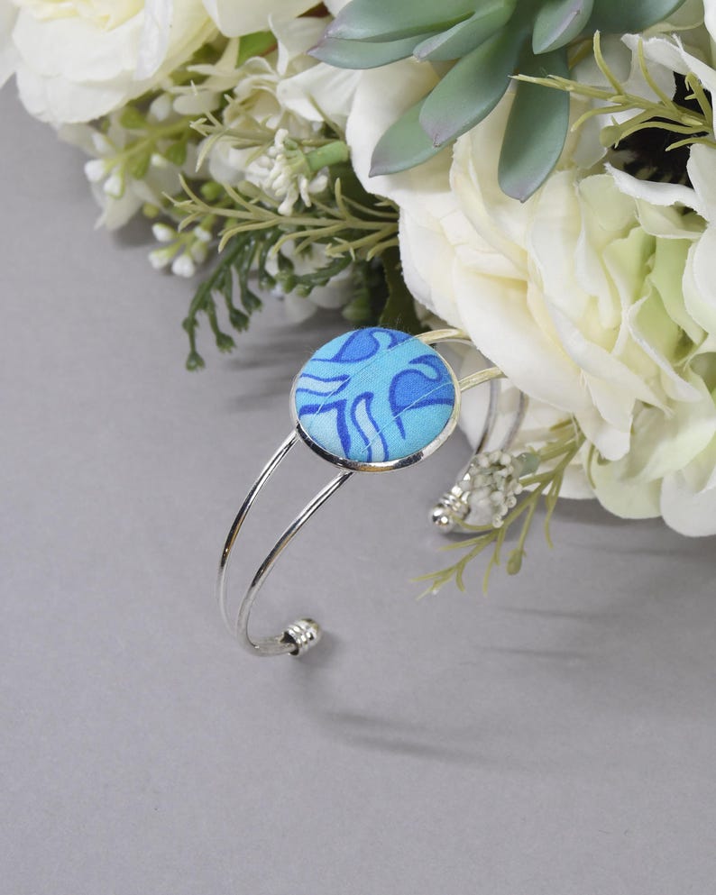 May include: A silver-toned metal cuff bracelet featuring a circular centerpiece covered in light and dark blue fabric with a swirling pattern. The bracelet has a double-wire band design that terminates in small, rounded decorative end caps.