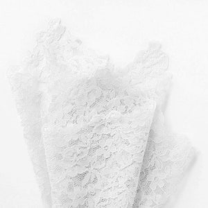 May include: A close-up view of white lace fabric, featuring a delicate floral pattern. The lace is layered and folded, creating a textured effect against a white background. The intricate design and soft color suggest a bridal or craft theme.