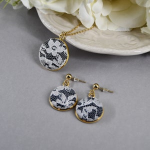 May include: A gold-toned necklace and earrings set. The pendant and earrings feature a circular design with white lace over a dark background, framed by a gold border. The jewelry is displayed on a gray surface.