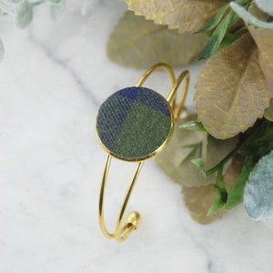 May include: A gold-toned metal cuff bracelet featuring a circular centerpiece inlaid with a fabric patch displaying a blue and olive green plaid pattern. The bracelet has a thin, double-wire band design that remains open at the back for adjustment.