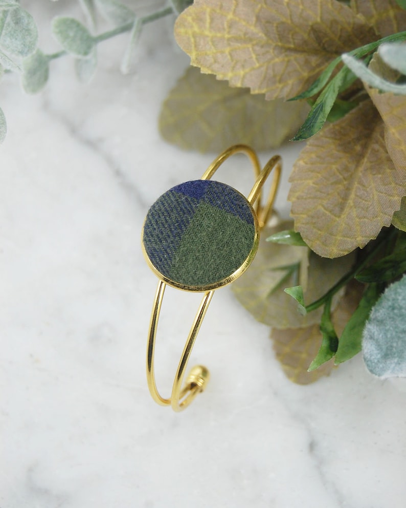 May include: A gold-toned metal cuff bracelet featuring a circular centerpiece inlaid with a fabric patch displaying a blue and olive green plaid pattern. The bracelet has a thin, double-wire band design that remains open at the back for adjustment.