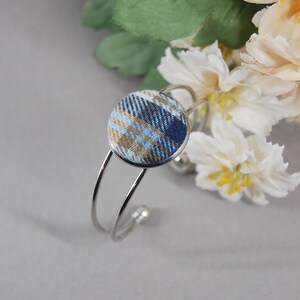 May include: A silver-toned metal cuff bracelet featuring a circular centerpiece covered in fabric with a blue, gold, and white plaid pattern. The bracelet has a double-wire band design with small rounded ends, showcasing a clean and polished metallic finish.
