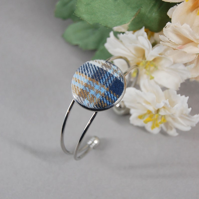 May include: A silver-toned metal cuff bracelet featuring a circular centerpiece covered in fabric with a blue, gold, and white plaid pattern. The bracelet has a double-wire band design with small rounded ends, showcasing a clean and polished metallic finish.