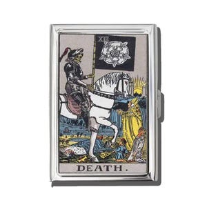 May include: A silver metal cigarette case featuring a tarot card design. The card depicts a skeletal figure on horseback holding a flag with a white rose. The word "DEATH" is printed at the bottom of the case. The artwork is in shades of black, white, yellow, blue, and red.