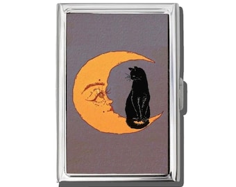 May include: A silver metal cigarette case featuring a whimsical design. The artwork depicts a black cat perched on an orange crescent moon with a face, set against a gray background. The case has a rectangular shape with a hinged closure.