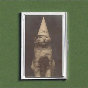Op de afbeelding: Een zilverkleurige metalen sigarettenetui in vintage-stijl met een sepia-kleurige afbeelding van een kat met een ezelsmuts. Het woord "DUNCE" staat op de muts. De etui staat tegen een groene achtergrond.