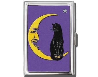 May include: A silver metal case with a purple background featuring a yellow crescent moon with a face, a black cat, and a star. The cat is sitting on the moon. The case has a hinged opening.