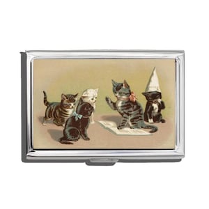 May include: A silver metal business card holder featuring a vintage illustration of several kittens. The kittens are in various poses, with one wearing a dunce cap. The background is a light tan color.
