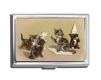 May include: A silver metal business card holder featuring a vintage illustration of several kittens. The kittens are in various poses, with one wearing a dunce cap. The background is a light tan color.