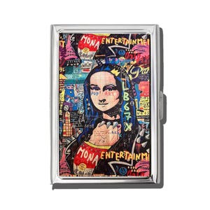 May include: A silver metal cigarette case featuring a pop art design of the Mona Lisa. The artwork includes vibrant colors, graffiti-style text, and the words "MONA ENTERTAINMENT" and "POP ART". The case has a hinged lid and a secure clasp.