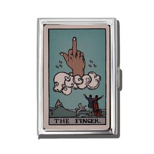 May include: A silver cigarette case with a tarot card design. The card shows a hand giving the middle finger from a cloud, with figures below. The text "THE FINGER" is at the bottom of the card.
