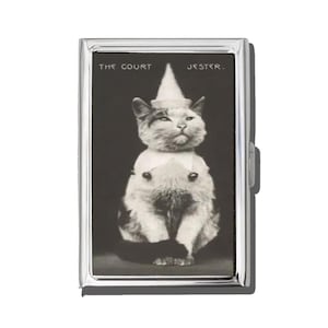 May include: A silver metal case featuring a black and white image of a cat dressed as a jester. The cat wears a white hat and collar, with the text "THE COURT JESTER." The case has a rectangular shape with rounded corners and a clasp closure.