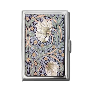 May include: A rectangular, silver-toned cigarette case with a floral design. The pattern features large, off-white flowers, green leaves, and small yellow blossoms against a dark blue background. The case has a clasp on the side.