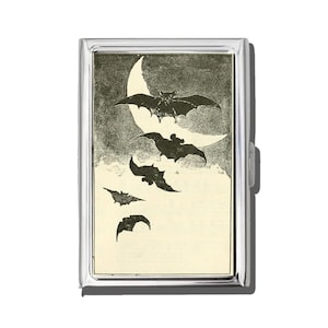 May include: A silver metal cigarette case featuring a vintage illustration of bats flying in front of a crescent moon. The artwork is in black and white, with a cream-colored background. The case has a hinged lid and a secure clasp.