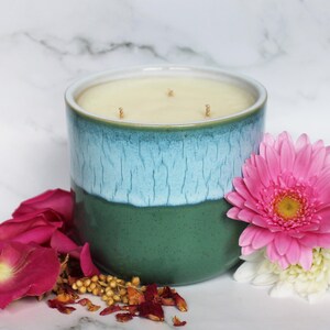 May include: A large, three-wick candle in a ceramic container. The container is two-toned, with a teal blue upper half and a green lower half. A pink gerbera daisy and rose petals are arranged near the candle.