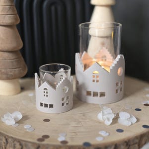 Op de afbeelding: Twee witte kandelaars met een huisvormig ontwerp. De kandelaars hebben uitgesneden ramen en deuren. De ene kandelaar is kleiner dan de andere. De kandelaars staan op een houten oppervlak met confetti en witte bloemen.