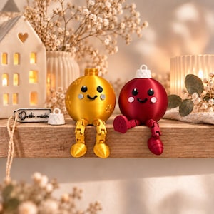 May include: Two spherical ornaments with articulated legs, one metallic gold and one deep red. Both feature smiling faces with black eyes and pink cheeks. A small white tag with the black text "Dreh mich" sits beside the gold ornament.
