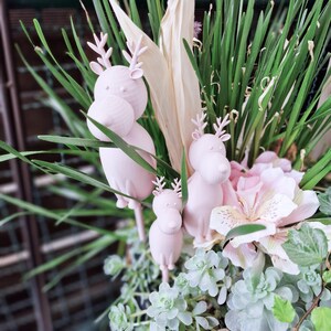 Puede incluir: Arreglo floral con figuras de renos rosas. El arreglo incluye hierba verde, flores rosa claro y varias plantas verdes y blancas. Los renos, de diferentes tamaños, añaden un toque caprichoso a la exhibición.