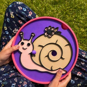 May include: A whimsical, layered wooden snail art piece. The snail features a pink body, a tan shell, and a black outline, set against a purple background within a pink and purple frame. A black flower and a purple sphere add detail.