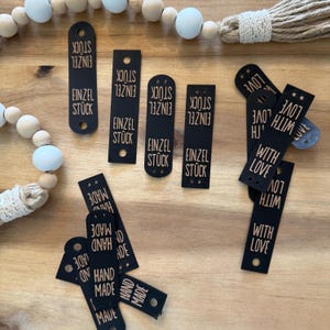 May include: A selection of black tags with gold lettering. The tags feature phrases such as "EINZEL STÜCK", "HAND MADE", and "WITH LOVE". Some tags have a rounded top, while others are rectangular. The tags are displayed on a wooden surface.