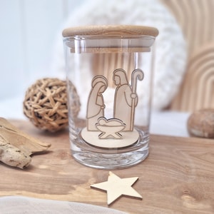 Può includere: Un barattolo di vetro trasparente con coperchio in legno, con un presepe in legno all'interno. La scena include le figure di Maria, Giuseppe e Gesù bambino. Una stella di legno è su una superficie di legno.