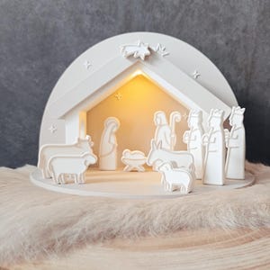 May include: A white, illuminated nativity scene with a star and figures of Mary, Joseph, the baby Jesus, the three wise men, and animals. The scene is set inside a white, arched structure with a warm, glowing interior.