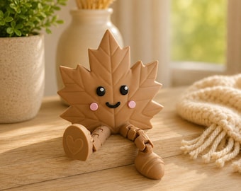 Puede incluir: Una figura de hoja de arce de color marrón claro con una cara sonriente, ojos negros y mejillas rosadas. La hoja tiene piernas articuladas con un pie en forma de corazón y el otro redondeado, diseñada para sentarse en un borde plano.