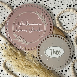 Könnte beinhalten: Zwei kreisförmige Holzschilder mit perforierten Rändern. Das größere altrosa Schild zeigt einen weißen Blattkranz und den Text "Willkommen kleines Wunder". Das kleinere beige Schild enthält den Namen "Theo" in einem weißen Oval.