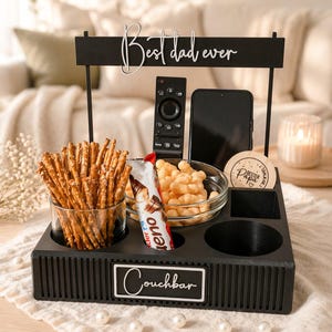 May include: A black wooden couch bar with the text "Couchbar" and "Best dad ever". It holds snacks, a remote, a phone, and a coaster. Snacks include pretzels, puffed snacks, and a candy bar.