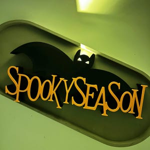 Könnte beinhalten: Ein Halloween-Schild mit einer schwarzen Fledermaus-Silhouette und den Worten "SPOOKY SEASON" in Orange. Das Schild befindet sich vor einem grünen Hintergrund und ist ein dekoratives Element.